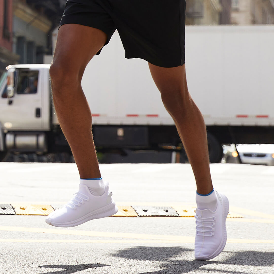 A person jogging outdoors on a city street wearing black athletic shorts, a white t-shirt, white sneakers, and carrying a gray shoulder bag.
