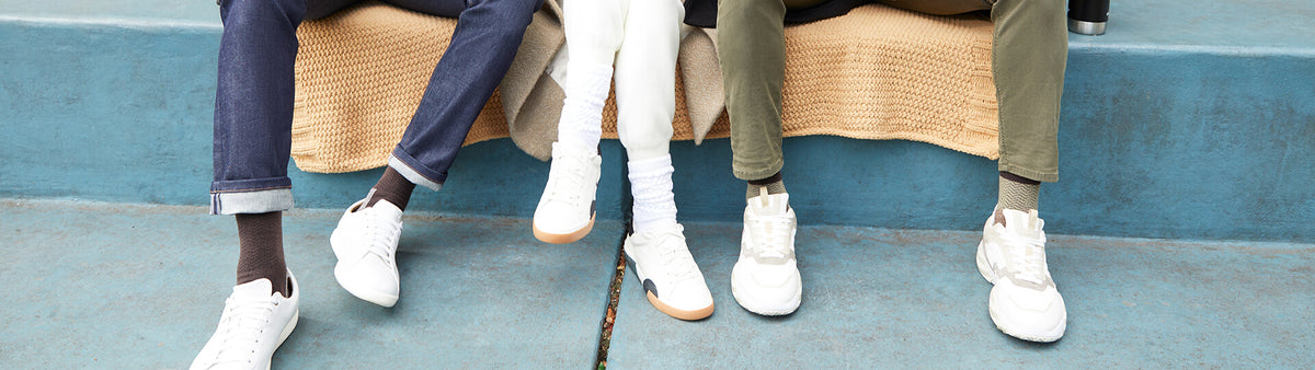 Three people sitting on a bench, shown from the knees down, wearing white sneakers. The scene features blue walls and a textured beige blanket.