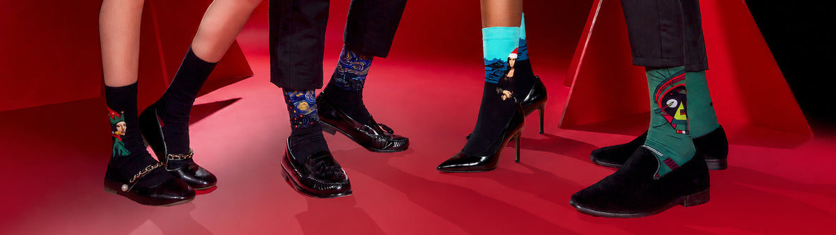 A close-up fashion shot showing multiple lower legs and feet wearing black shoes and colorful illustrated socks against a red studio backdrop.