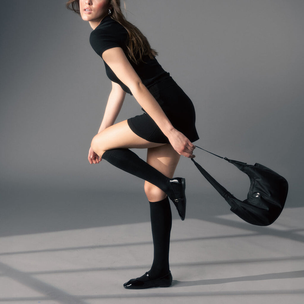 Woman in black outfit with knee-high socks and a black handbag on a gray background