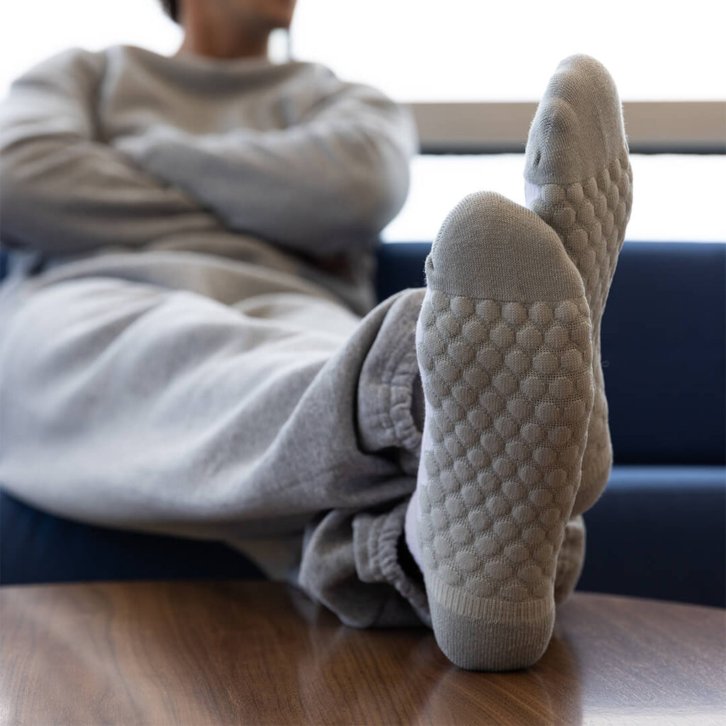 A person reclining indoors with their legs stretched out, wearing light gray knit socks with a raised bubble texture on the soles. The scene is shot from a low angle, with the person and a blue chair blurred in the background.