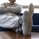 A person reclining indoors with their legs stretched out, wearing light gray knit socks with a raised bubble texture on the soles. The scene is shot from a low angle, with the person and a blue chair blurred in the background. thumbnail
