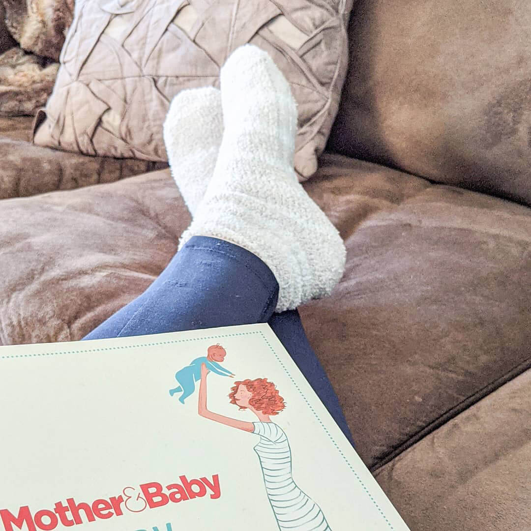 Person wearing white socks and blue leggings with a 'Mother & Baby' magazine on a brown couch.