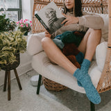 Person sitting on a white chair wearing fuzzy socks reading a book with plants and a wicker chair in the background. thumbnail