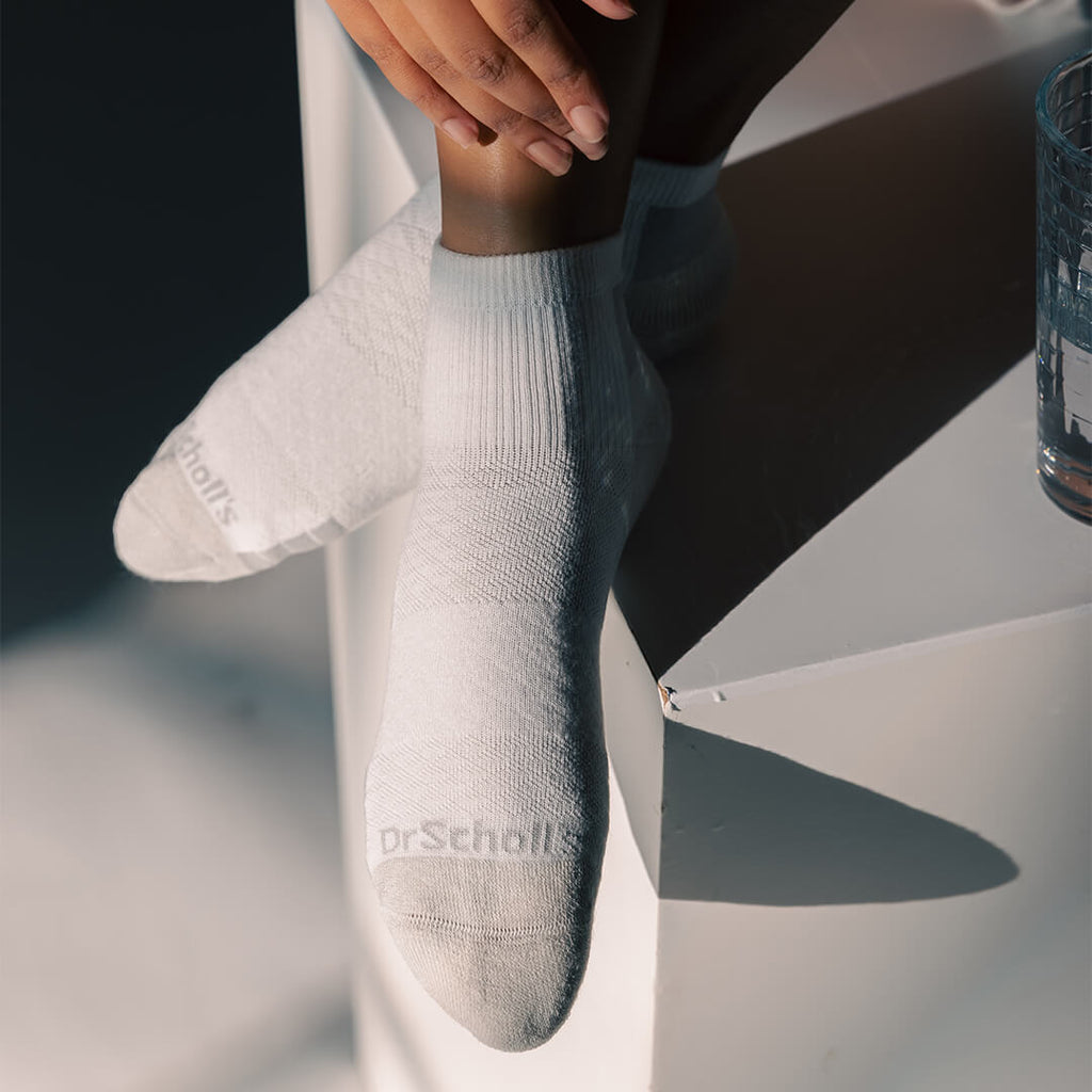 A person wearing white ankle socks with a subtle textured pattern and the brand name 'Dr. Scholl's' printed on the toes, posed on a white geometric surface with dramatic shadows.