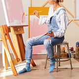 A woman sitting on a metal stool in an art studio, wearing blue denim overalls and striped blue-and-white long-sleeve shirt, holding paintbrushes. She is positioned next to a wooden easel with pink and orange paint on the canvas, with other art supplies on a stool behind her. thumbnail