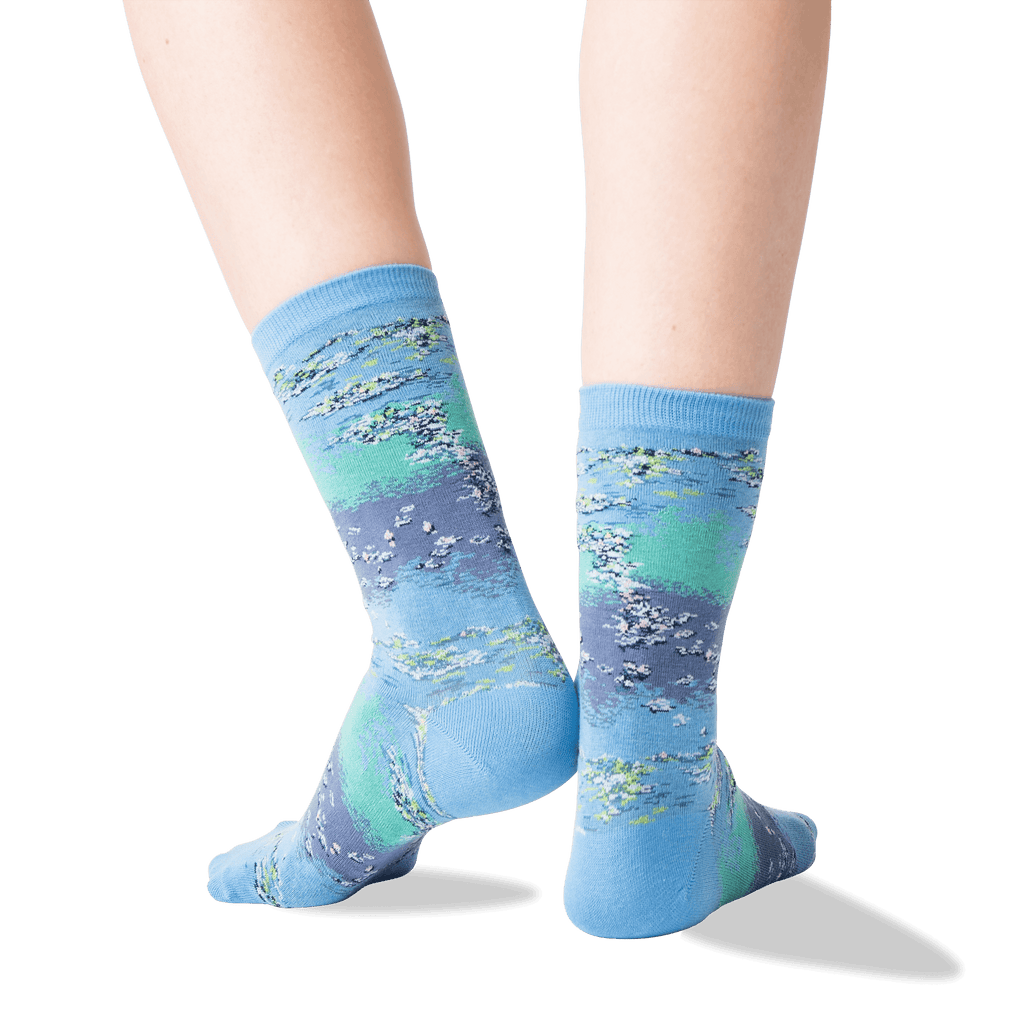 A pair of crew socks worn on feet, featuring an abstract landscape-like print in shades of light blue, teal, and lavender-gray with white and gray brushstroke details. The background is a clean white studio backdrop.