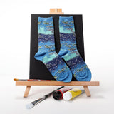 A pair of crew socks with a blue and teal watercolor-style floral pattern, displayed on a black backdrop stand on a wooden artist easel with paint tubes and brushes. thumbnail