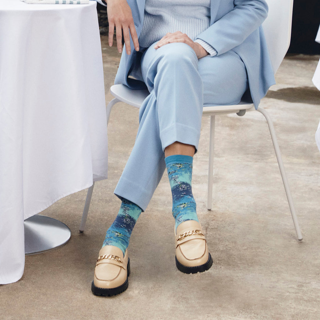 A person seated on a white chair wearing light blue tailored clothing and patterned blue socks with beige loafers, photographed in a real indoor setting with a table and floor visible.