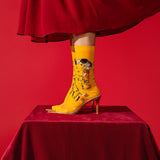 A person's leg wearing a bright yellow pointed-toe high-heeled boot with a surreal collage print, standing on a red velvet-covered pedestal against a solid red background. thumbnail