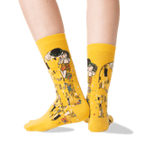 A pair of bright yellow crew socks with a Gustav Klimt 'The Kiss' graphic print, shown on a model's feet against a clean white background.