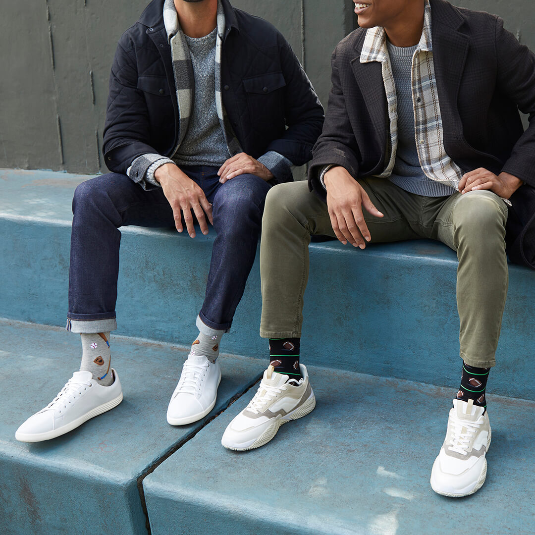 Two men sitting on blue concrete steps wearing white low-top sneakers and HOTSOX sports-themed novelty crew socks. They are dressed in casual jackets, shirts, and jeans or chinos.