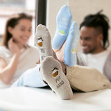 A person lying down with their feet raised, wearing gray socks printed with a beer mug and the words 'POT HEAD' and light blue socks printed with a beer mug and the words 'I LOVE YOU SO MATCHA'. Two blurred people are visible in the background.
