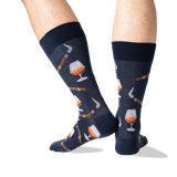 A pair of navy blue crew socks with a pattern of whiskey glasses, cigars, and smoking pipes, shown on a person's legs against a white background.