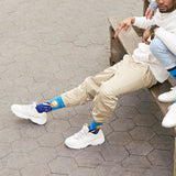 A man sitting on a wooden bench outdoors on a gray, hexagonal-paved surface, wearing beige cargo pants, white sneakers, and bright blue crew socks with Van Gogh's "Self Portrait" knit into the design. thumbnail
