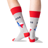 A person's legs wearing heather gray crew socks with red cuffs, heels, and toes, featuring a graphic of the state of Texas with the Texas flag and the word 'TEXAS'.