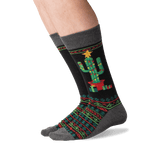 A pair of crew socks worn on a person's feet, featuring a black background with a green cactus in a red pot, wrapped in festive red and green geometric patterns, and topped with a yellow star. thumbnail
