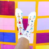 White crew socks with a colorful pattern of cocktail glasses, shown on a person's feet against a vibrant, multicolored tiled wall.