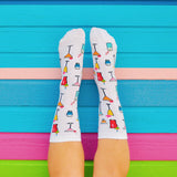 A pair of white crew socks with a colorful cocktail glass pattern, worn on feet against a bright blue wooden background with pink and green stripes. thumbnail