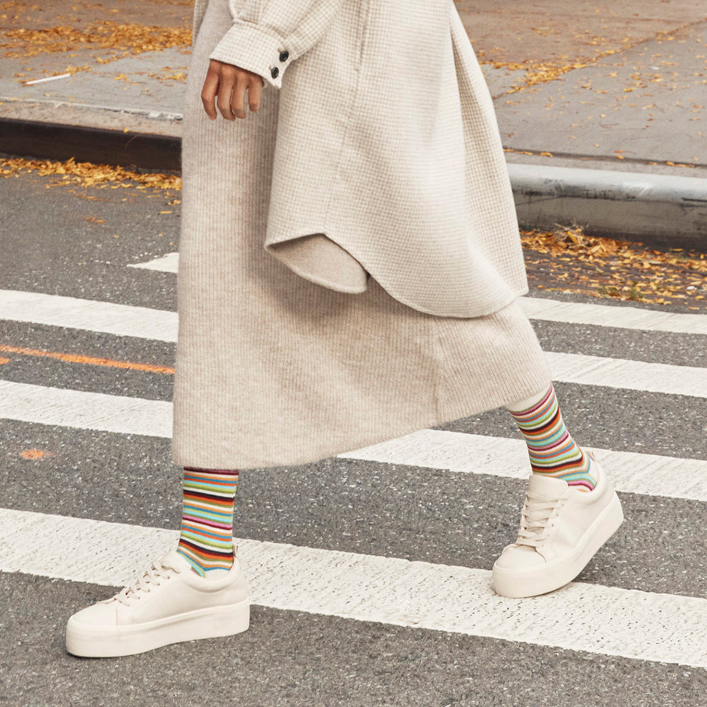 A person walking across a crosswalk wearing a long cream knit dress, a light beige cardigan, colorful striped socks, and white platform sneakers.