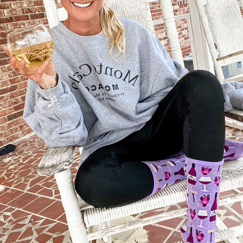 Person sitting on a porch holding a glass of wine, wearing a gray sweatshirt and colorful socks with a wine pattern.