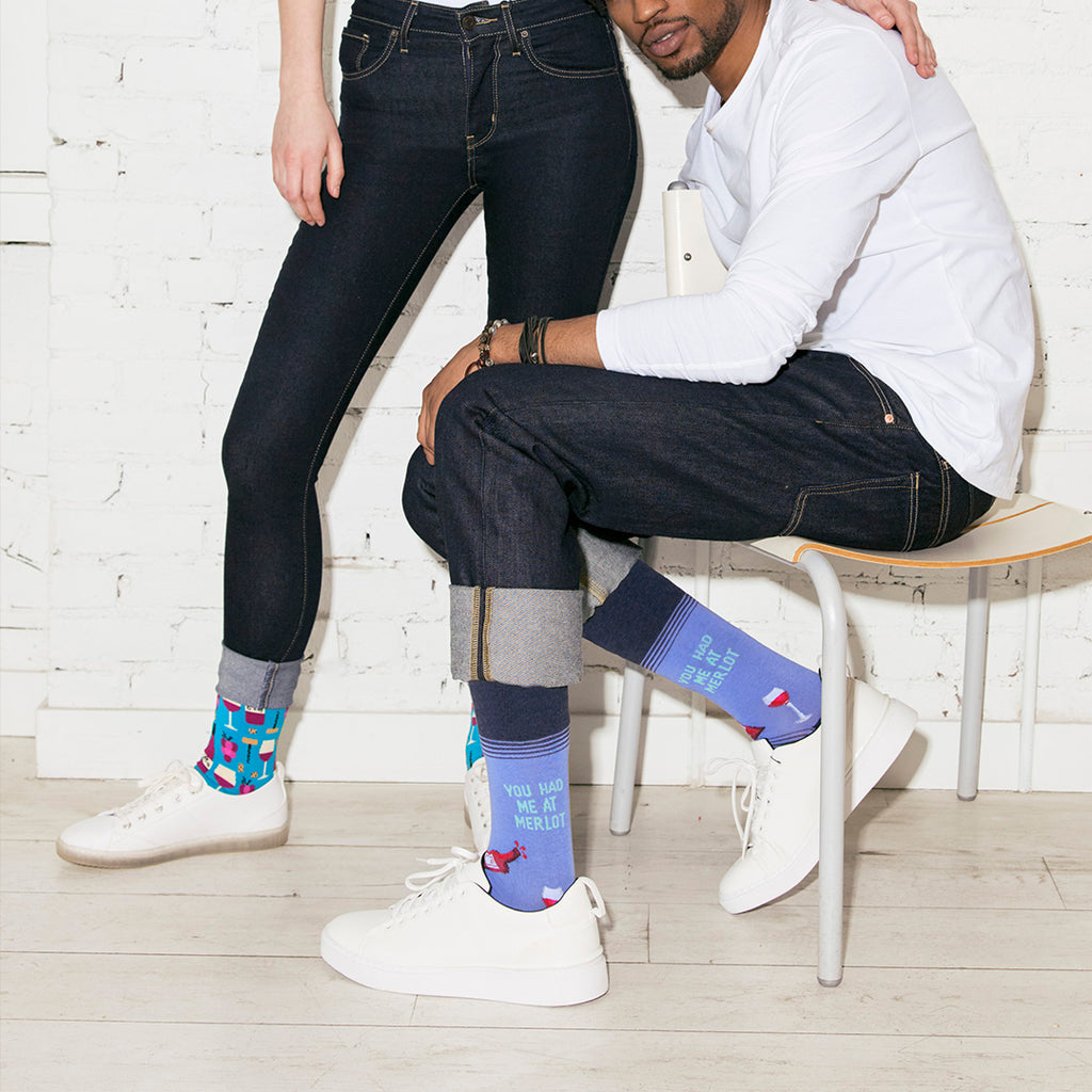A man and a woman seated indoors wearing dark jeans and white sneakers, with the man's legs crossed over a white chair. The man's socks are blue with a red wine bottle and the text 'YOU HAD ME AT MERLOT, while the woman's socks are multicolored with a geometric wine  pattern.