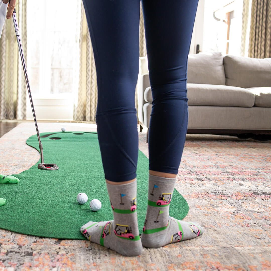 Person playing mini golf indoors with a green mat and golf ball on a patterned rug.