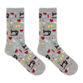 A pair of heather gray crew socks with an all-over sewing-themed pattern featuring black sewing machines, red hearts, yellow spools, scissors, and other colorful thread icons, shown against a plain white background.