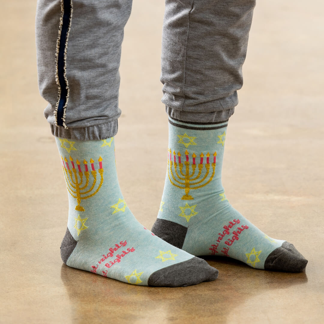 A person wearing light blue crew socks with a yellow menorah, pink candles, and pink text, paired with gray sweatpants, standing on a wooden floor.