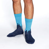A person's legs wearing bright blue crew socks with a dark navy blue dripping graphic design on the feet, photographed against a seamless white background. thumbnail