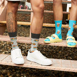 Two people standing on a metal walkway over gravel, wearing patterned crew socks. One person wears gray socks with a brown and orange sun glasses print, paired with white sneakers.