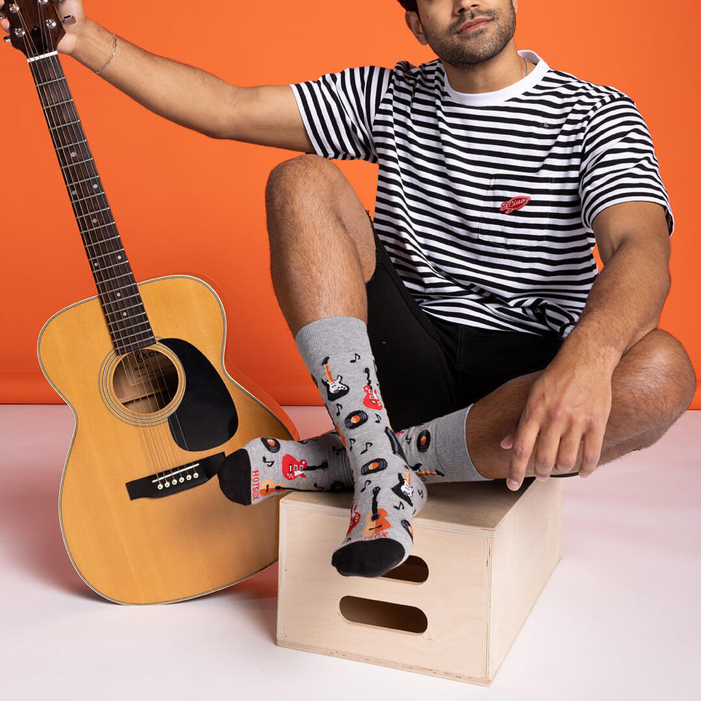 Person sitting with a guitar, wearing striped shirt and colorful socks against an orange background