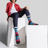 A model wearing colorful striped crew socks, a red button-up shirt, and dark jeans sits on white geometric display blocks against a light gray studio background.