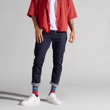 A model wearing dark blue slim-fit jeans with rolled cuffs, styled with a red striped short-sleeve button-up shirt, a white t-shirt, colorful striped socks, and white sneakers, set against a plain light gray studio background.