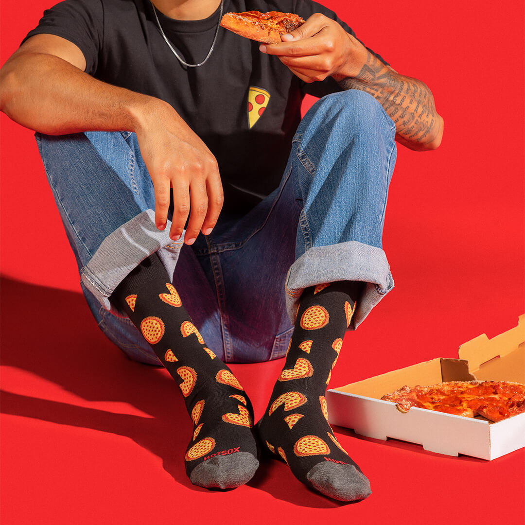 A person sitting on a bright red floor wearing black socks with a repeating pizza slice pattern, holding a slice of pizza. A pizza box is open on the floor beside them.