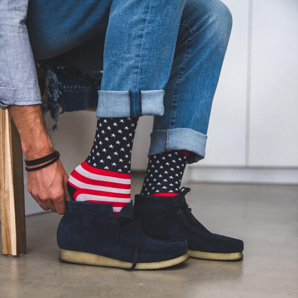 A person wearing blue jeans and dark navy suede boots, with one hand adjusting patriotic socks that feature a white star pattern on a black background and red-and-white stripes near the top.