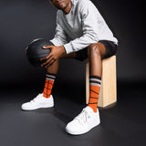 Person sitting on a wooden block holding a basketball, wearing gray sweatshirt, black shorts, white sneakers, and orange socks with black stripes.