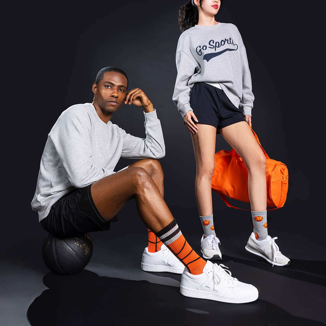 Two people wearing gray sweatshirts with 'Go Sports' text, black shorts, and orange socks on a dark background.