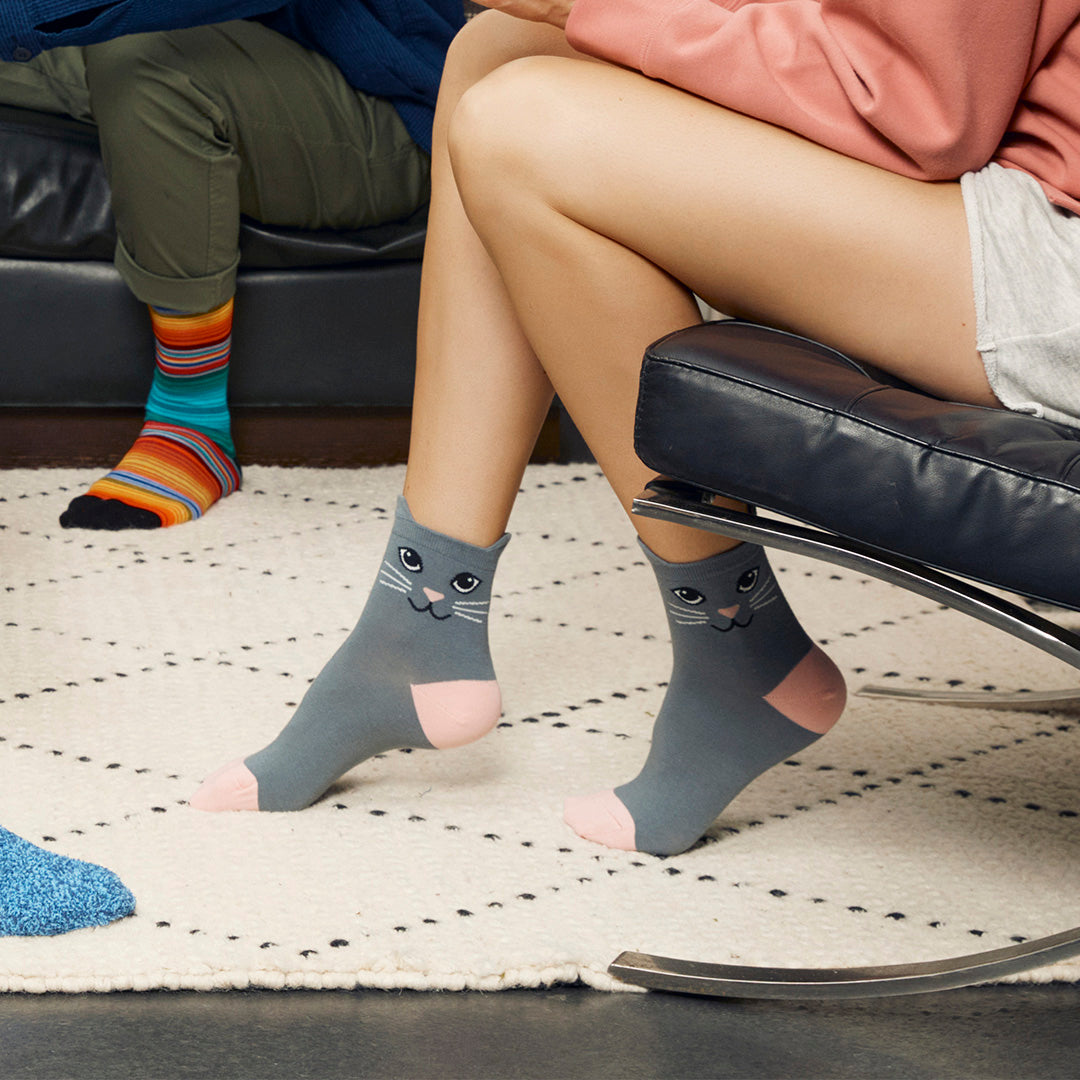 A person sitting on a black chair wearing gray socks with a cat face design and pink toes, with another person's legs visible in the background wearing colorful striped socks in a home setting.