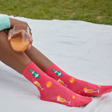 A person sitting outdoors on a white blanket holds a glass of orange drink while wearing bright pink crew socks printed with cocktail and citrus motifs. Grass is visible in the background.