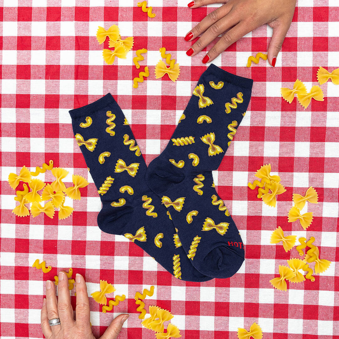 Navy blue socks with yellow pasta patterns on a red and white checkered tablecloth with scattered yellow pasta.