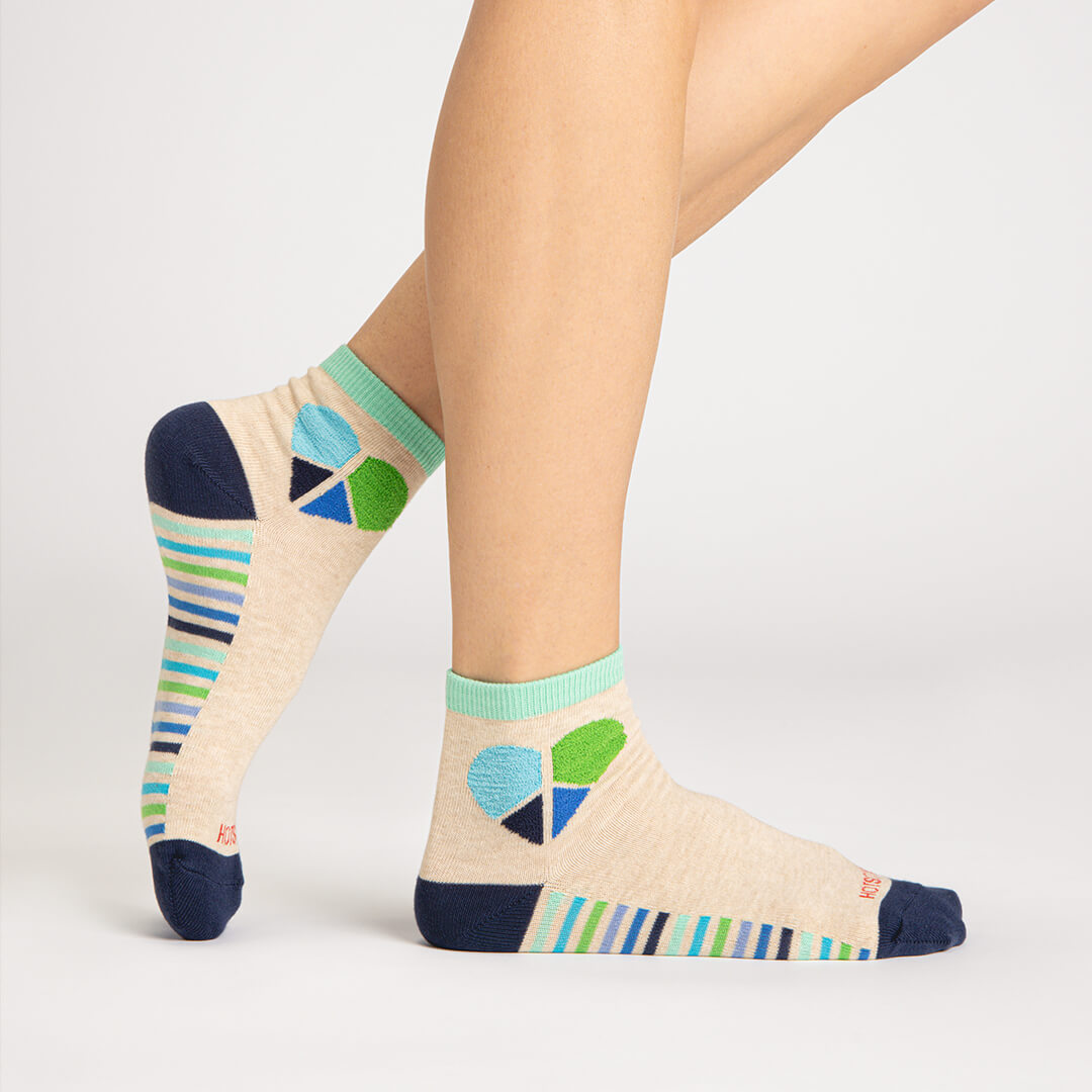 A pair of ankle socks worn on a model's feet, shown against a clean light gray studio background. The socks are heather beige with a colorful geometric heart design in blue, green, and navy on the ankle, striped bands in blue, green, and white on the sides, and solid navy heels, toes, and cuffs.