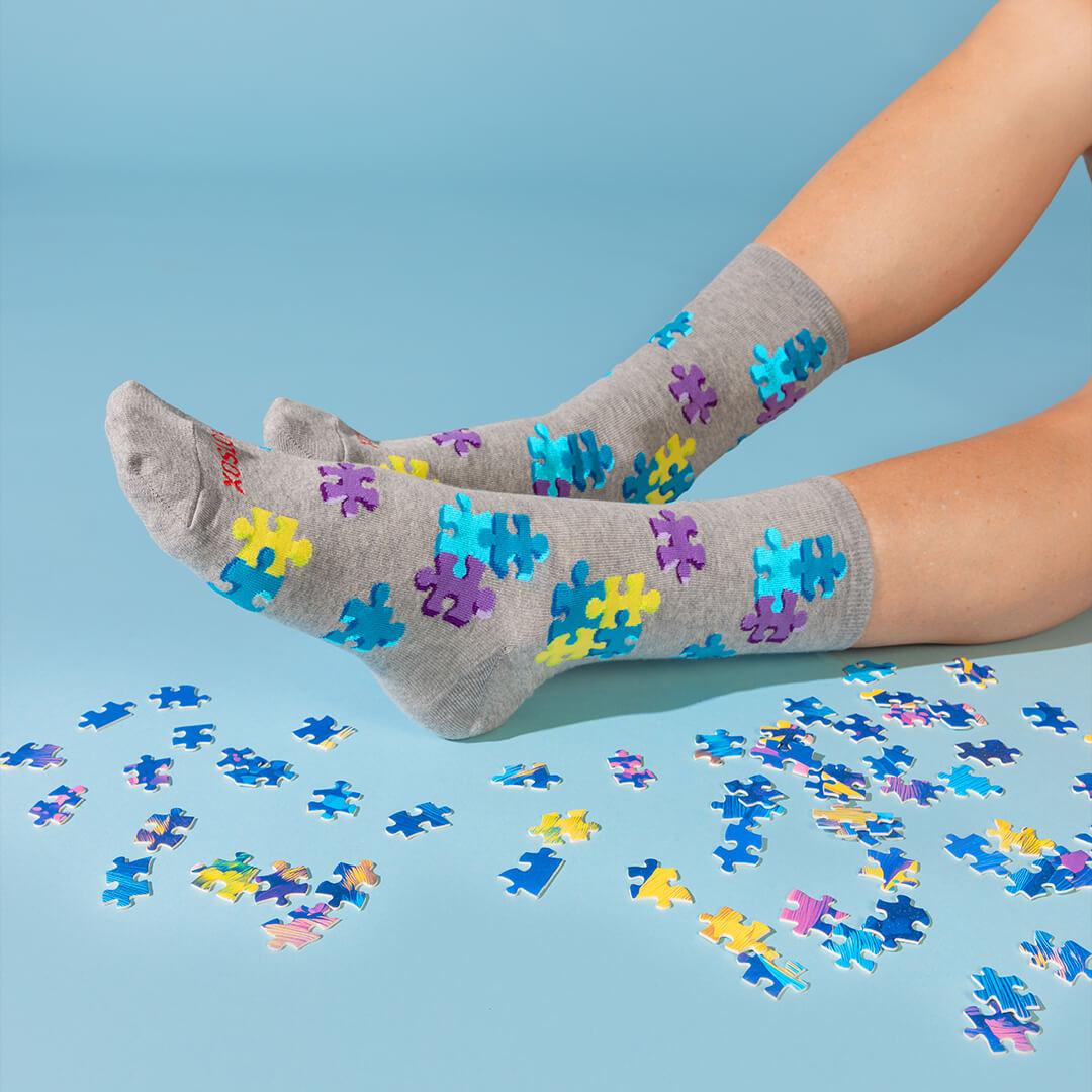 A pair of gray crew socks with colorful jigsaw puzzle-piece patterns, worn on feet with scattered puzzle pieces on a light blue background.