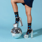A person wearing black crew socks with a colorful space-themed graphic and silver high heels, posed with one foot on a mirrored disco ball against a light blue studio background. thumbnail