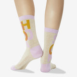 A pair of crew socks worn on a model's legs, featuring a light beige heathered base with lavender cuffs, heels, and toes, and a graphic print of yellow and lavender letters. thumbnail
