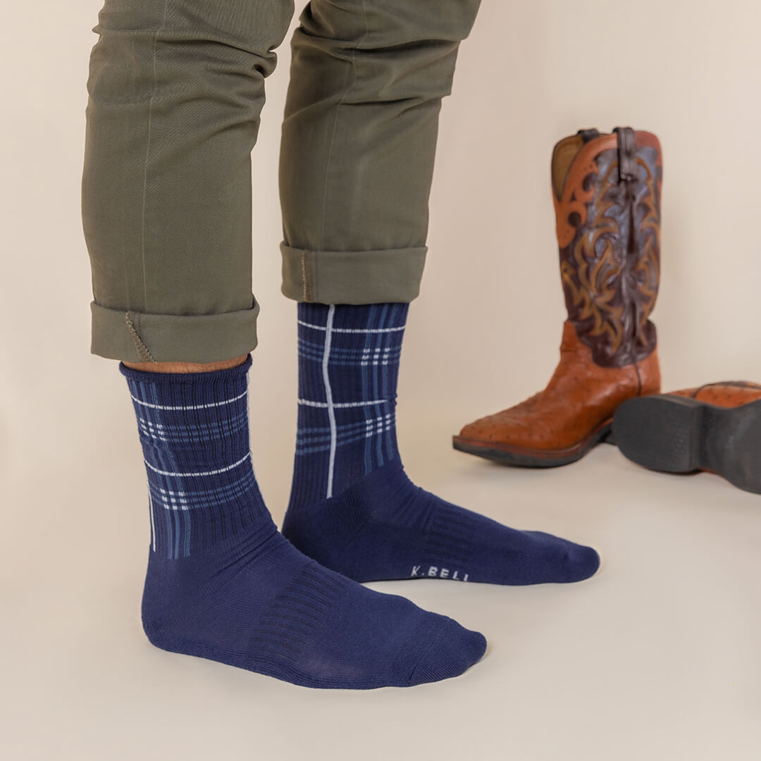 Person wearing blue plaid socks with brown cowboy boots on a neutral background