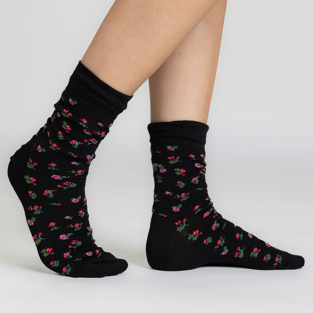 A pair of black crew socks with a small pink and green floral pattern, shown on a model's feet against a light gray studio background.