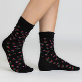 A pair of black crew socks with a small pink and green floral pattern, shown on a model's feet against a light gray studio background.