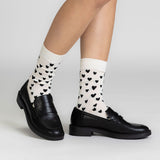 A person's legs wearing cream-colored crew socks with a black heart pattern, paired with black leather penny loafers, shown against a light gray studio background.