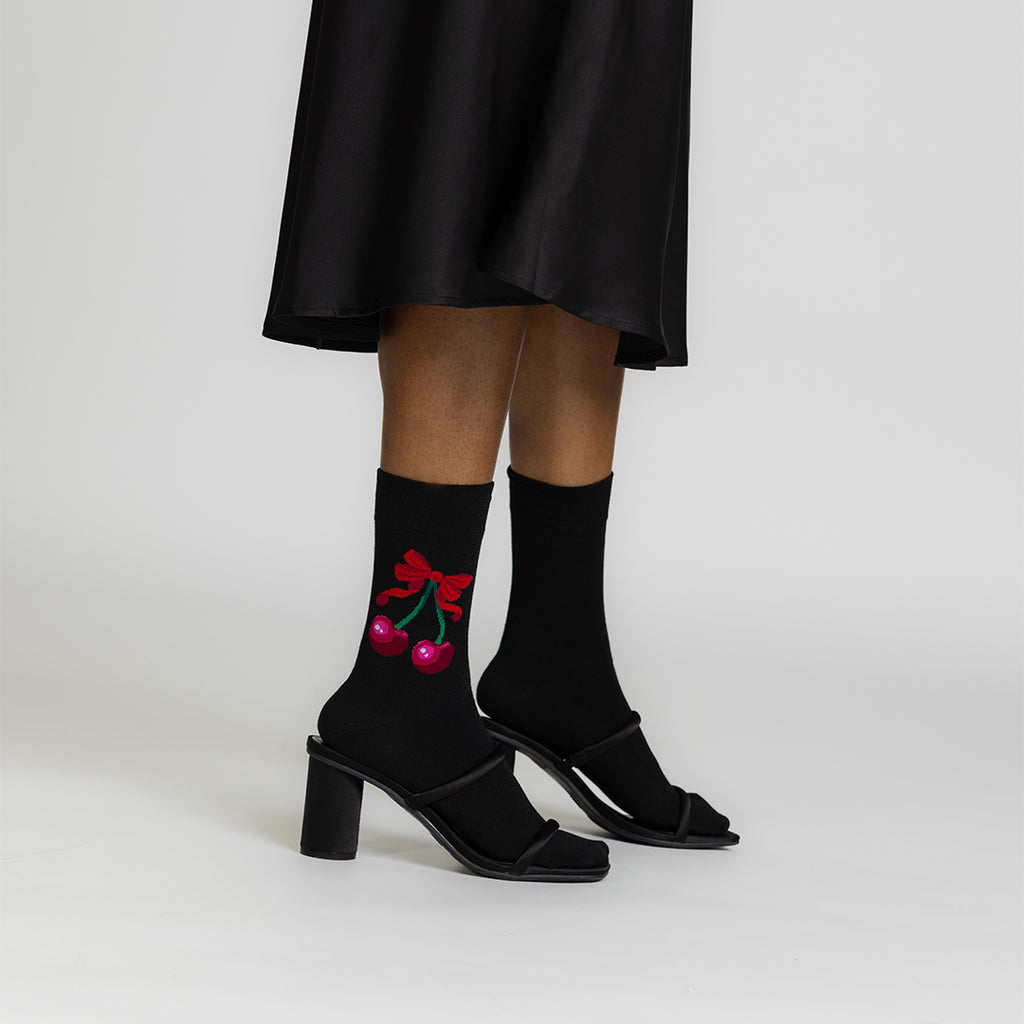 A person wearing black mid-heel sandals and black crew socks with a red cherry and bow graphic, shown from the knees down against a light gray studio background.